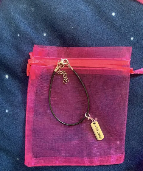 A black cord bracelet with a silver tag engraved willPower, on a pink organza pouch.