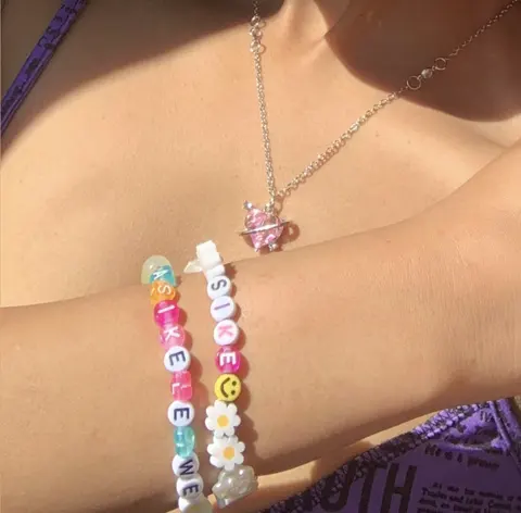 Two letter bead bracelets on a wrist, spelling ASIKELELWE and SIKE, worn with a pink heart pendant necklace.