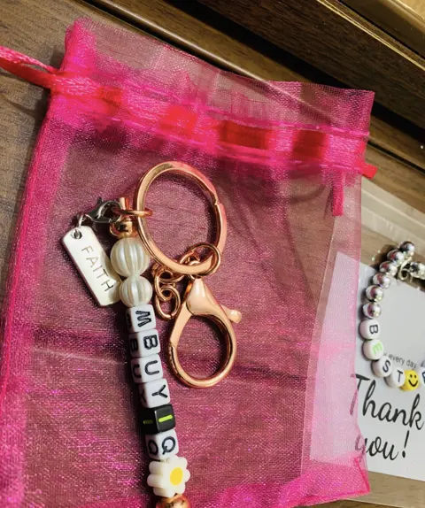 A rose gold key chain with a FAITH tag, pearl beads, a daisy and letter beads spelling MBUYI, on a pink organza pouch.