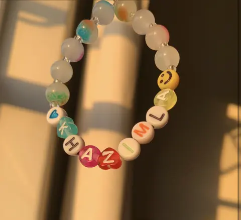 A bracelet of frosted pastel beads, a smiley bead and letter beads spelling KHAZIMLA, hanging in warm sunlight.