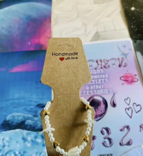 A pearl bead bracelet with gold heart beads and white flower charms on a kraft card that says Handmade with love.