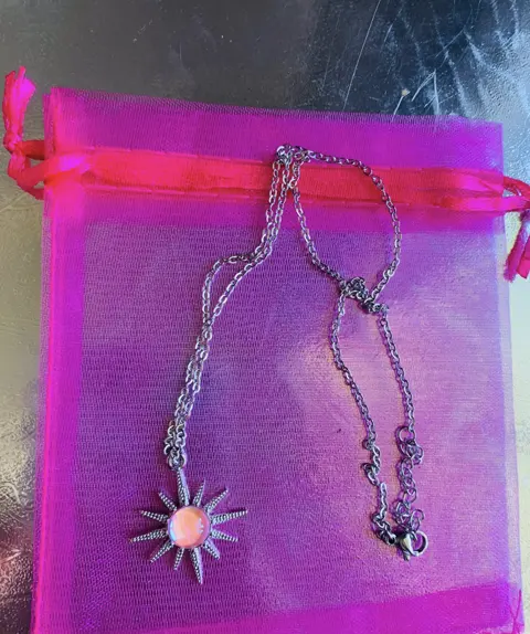 A silver chain necklace with a sunburst pendant and a pink glass centre, on a pink organza pouch.