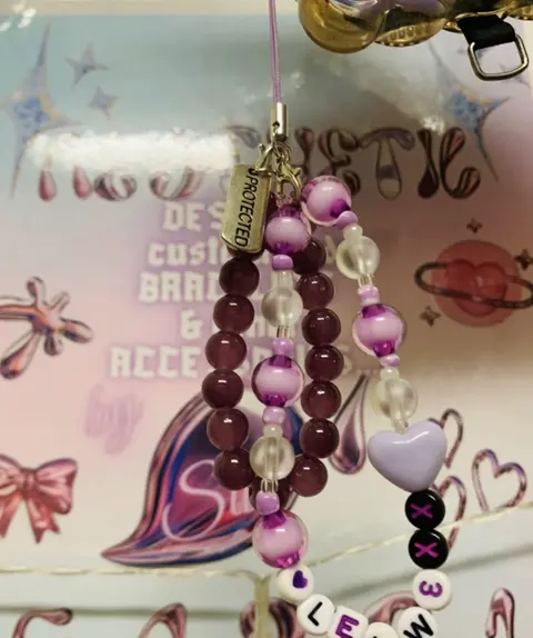A beaded phone strap in plum and lilac with a heart bead, a PROTECTED tag and letter beads spelling LELW.